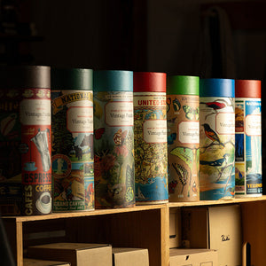 A row of vintage-style illustrated puzzles  in assorted styles displayed on a wooden shelf, with cardboard boxes and mugs below in a dimly lit setting.